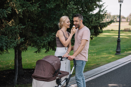 wife feeding husband with ice cream near baby carriage in park