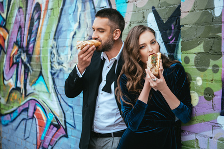 portrait of couple in luxury clothing eating hot dogs near wall with graffiti on streetの写真素材