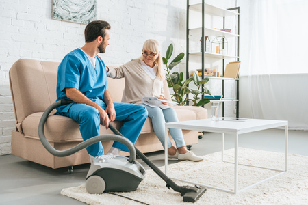 smiling senior woman sitting on couch and looking at social worker with vacuum cleanerの写真素材