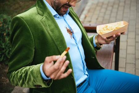 partial view of man in green velvet jacket with ketchup on shirt and french fries in hands sitting on bench on streetの写真素材