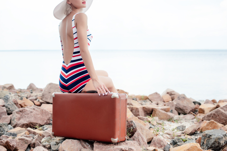 cropped view of stylish woman in striped swimsuit sitting on luggage on rocky beachの写真素材
