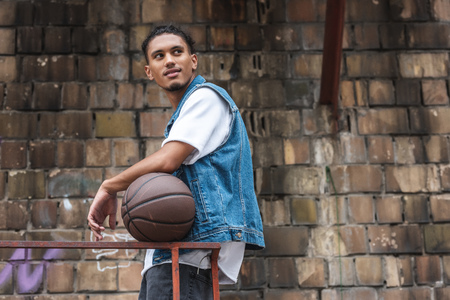 stylish mixed race man standing with basketball ball and looking away at streetの写真素材