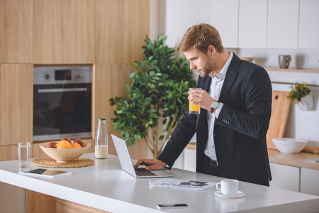 focused businessman drinking fresh juice using laptop at kitchen table during breakfastの写真素材