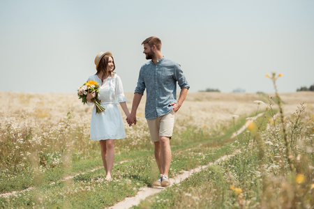 smiling woman in white dress with bouquet of wild flowers walking together with boyfriend in fieldの写真素材