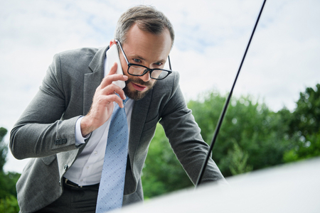 handsome businessman talking by smartphone and looking in open car hoodの写真素材
