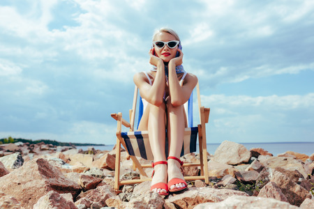attractive stylish woman in retro swimsuit relaxing in beach chair on rocky shoreの写真素材