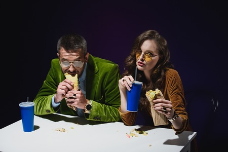portrait of couple in luxury velvet clothing with shawarma and soda drinks sitting at table with dark background behindの写真素材