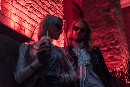 young stylish couple in fashionable clothes standing together under red light on night streetの写真素材