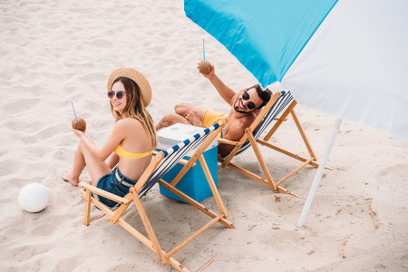 high angle view of happy young couple with coconut cocktails relaxing in sun loungers on sandy beachの写真素材