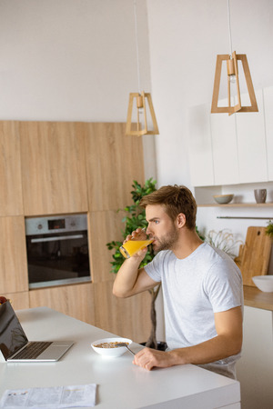 young man drinking fresh juice at table with plate of flakes and laptop at kitchenの写真素材