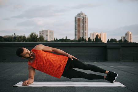 handsome sportsman doing side plank on yoga mat on roofの写真素材