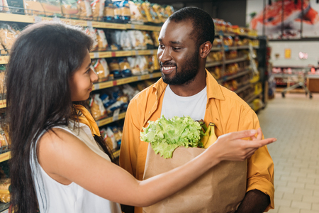 young african american woman pointing to boyfriend with paper bag with food in supermarketの写真素材
