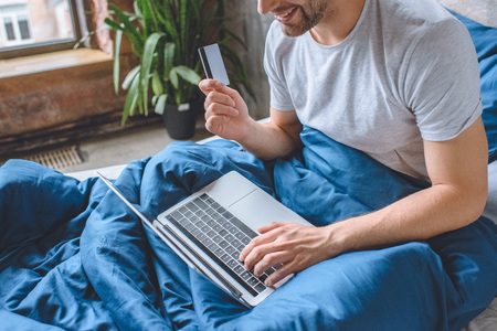 cropped image of young man in bed with credit card doing online shopping on laptopの写真素材