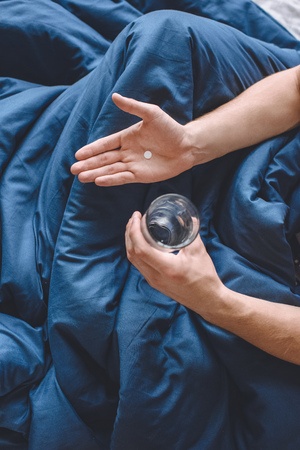 cropped image of young man with headache holding pill and glass of water in bed at homeの写真素材
