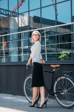 young businesswoman in stylish clothing with retro bicycle standing on streetの写真素材