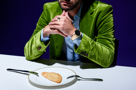 cropped shot of man in stylish green velvet jacket sitting at table with cheburek on plate with blue background behindの写真素材