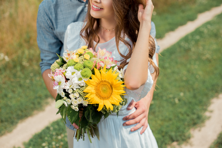 partial view of man hugging smiling girlfriend with bouquet of wild flowers in summer fieldの写真素材