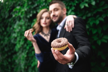 selective focus of fashionable couple with chocolate doughnuts with green foliage behindの写真素材