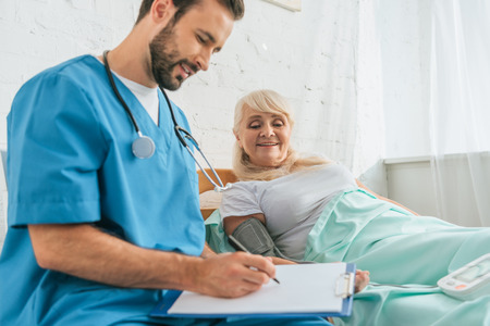 smiling male nurse writing on clipboard while measuring blood pressure to senior woman lying in bedの写真素材