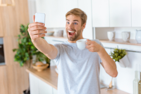 happy young man with coffee cup taking selfie on smartphone at kitchenの写真素材