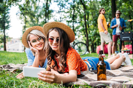 selective focus of smiling friends taking selfie on smartphone while resting on blanket during bbq in parkの写真素材