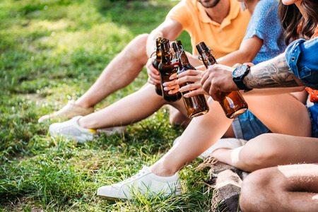 cropped shot of clinking bottles of beer while resting on green grass in parkの写真素材