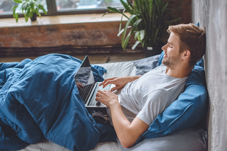 smiling man in bed using laptop with booking website on screenの写真素材