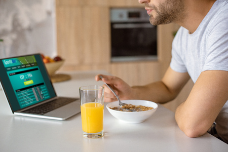 cropped image of man eating flakes with milk at kitchen table with fresh juice in glass and laptop with sportsbet on screenの写真素材