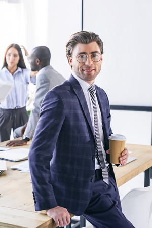 selective focus of caucasian businessman with coffee to go and multiracial colleagues behind in officeの写真素材