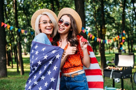 portrait of happy women in hats and sunglasses with american flag in park