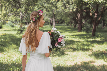 back view of tender young bride holding wedding bouquet and walking in parkの写真素材