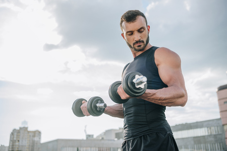 handsome sportsman training with dumbbells on roof and looking at bicepsの写真素材