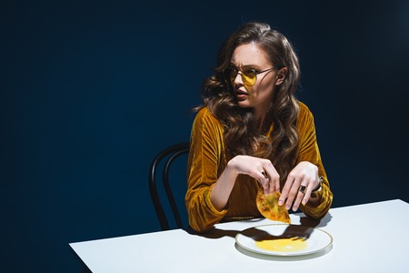 fashionable woman with unhealthy meat pastry sitting at table with blue backdropの写真素材