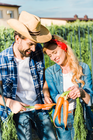 smiling couple of farmers holding organic carrots in field at farmの写真素材
