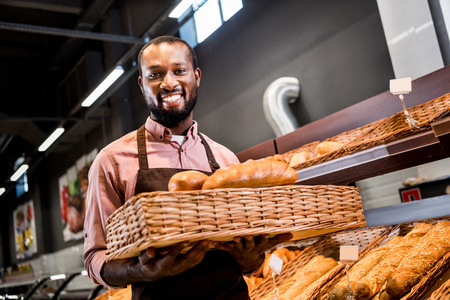 selective focus of smiling male african american shop assistant in apron holding loaves of bread in grocery storeの写真素材