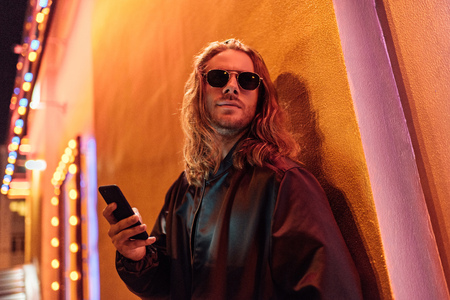 bottom view of attractive young man in leather jacket and sunglasses using smartphone and looking away on street at night under yellow lightの写真素材
