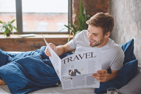 smiling young man reading travel newspaper in bed at homeの写真素材