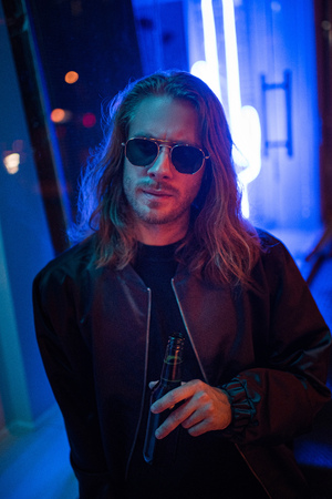handsome young man in leather jacket and sunglasses with bottle of beer on street at night under...