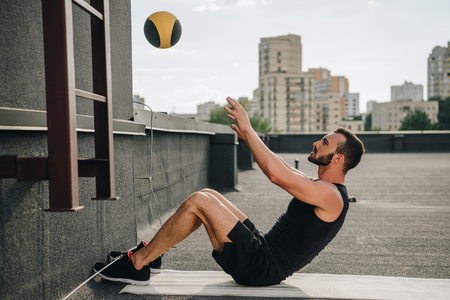side view of handsome sportsman doing sit ups with medicine ball on yoga mat on roofの写真素材