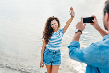 man taking photo of attractive waving girlfriend near sea on smartphone