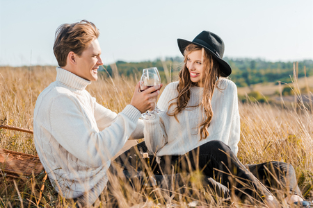 young couple clinking with glasses of red wine and looking at each other during picnic on meadowの写真素材