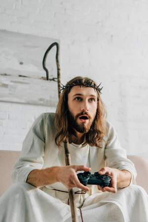 emotional Jesus with wooden staff playing video game by joystick on sofa at homeの写真素材