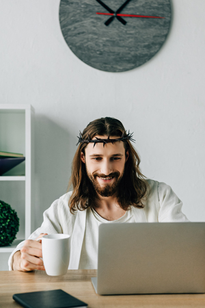 cheerful Jesus in crown of thorns with coffee cup using laptop at table in modern officeの写真素材