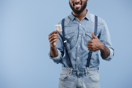 cropped image of smiling african american man showing thumb up to condom isolated on blueの写真素材