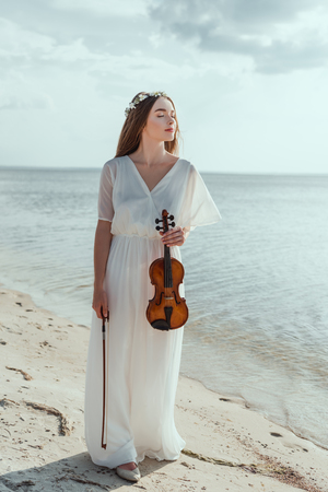 attractive elegant girl in white dress holding violin on seashoreの写真素材