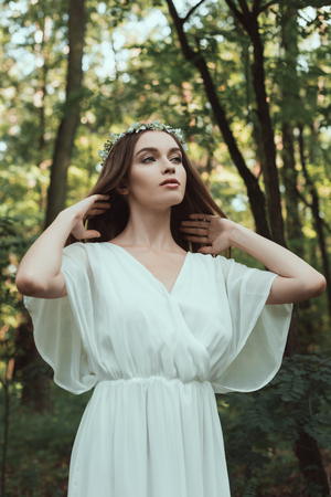 young tender woman posing in white dress and floral wreath in forestの写真素材