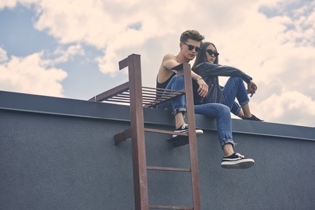 stylish multicultural couple in sunglasses sitting on roof with ladderの写真素材