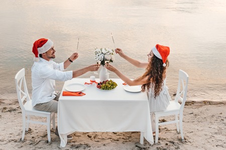 young couple in santa hats holding sparklers and toasting with champagne glasses while celebrating christmas on beachの写真素材