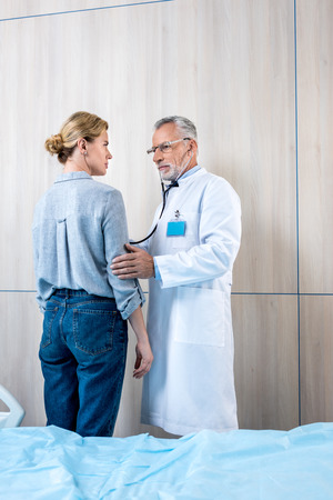 confident mature male doctor examining female patient by stethoscope in hospital roomの写真素材
