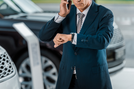 cropped shot of adult businessman talking by phone at car dealership salon and looking at wrist watchの写真素材